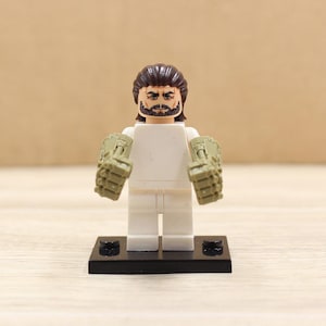 May include: A white Lego figure with brown hair and a black beard, wearing olive green gauntlets. The figure stands on a black base against a neutral background. A collectible toy.