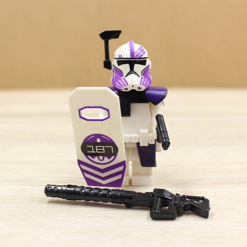 May include: A white and purple action figure of a soldier with a helmet, shoulder armor, and a shield with the text "187". A black rifle is in front of the figure. The figure is a toy.