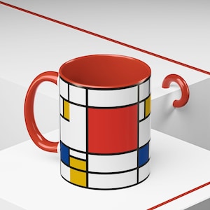 May include: A red and white ceramic mug with a geometric design in red, yellow, and blue. The mug has a black outline around the design.
