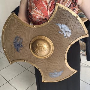 May include: A brown, wood-textured shield with a gold-colored rim and central dome. The shield features a raised gold-colored triskelion design on the center boss and three distinct, stylized animal-like silhouettes in blue and white.