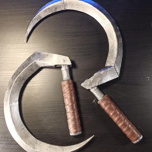 Peut inclure: Deux faucilles en métal argenté avec des manches en bois brun. Les faucilles sont disposées en cercle, les manches pointant vers l'extérieur.