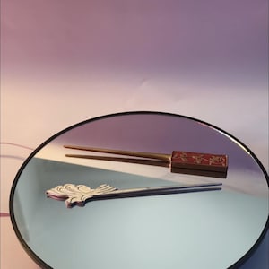 Peut inclure: Deux épingles à cheveux décoratives sont présentées sur une surface réfléchissante. L'une est dorée avec un accent rectangulaire rouge. L'autre est argentée avec un motif décoratif en forme de vague. Le fond est un dégradé de violet et de blanc.