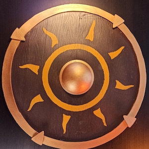 Puede incluir: Un escudo redondo de madera con un borde dorado y un diseño de sol pintado en amarillo. El escudo tiene un pequeño jefe redondo de color dorado en el centro.