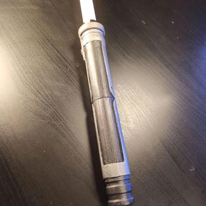 以下が含まれることがあります： テクスチャ加工されたグリップを持つ黒と銀のライトセーバーの柄。柄は中央に銀色のバンドがあり、先端に黒いキャップが付いています。