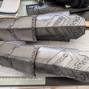 May include: A pair of metallic silver arm guards featuring intricate embossed geometric patterns and knotwork designs. The guards are positioned horizontally, with a wooden ruler visible in the background showing measurements in inches.