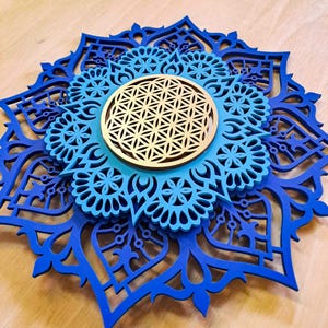 Op de afbeelding: Een blauwe en gouden mandala wandkunst met een geometrisch patroon in het midden. De mandala is gemaakt van hout en heeft een gelaagd ontwerp.