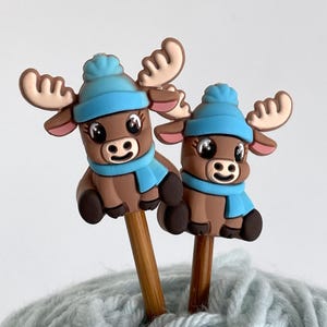 Könnte beinhalten: Zwei braune Elch-Stricknadelaufsätze. Jeder Elch trägt eine blaue Mütze und einen Schal. Die Aufsätze befinden sich auf Holzstricknadeln und ruhen auf einem hellblauen Wollknäuel.