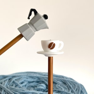 Könnte beinhalten: Eine Miniatur-Kaffeemaschine in Grau und eine weiße Kaffeetasse mit einem braunen Kaffeebohnen-Design sind an Stricknadeln aus Holz befestigt. Im Hintergrund befindet sich ein Knäuel hellblauer Wolle.