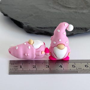 May include: Two pink gnome figurines with white polka dots and a ruler. One gnome is standing, the other is lying down. The standing gnome has a white beard and a pink hat with a white pom-pom. The ruler shows measurements in centimeters.