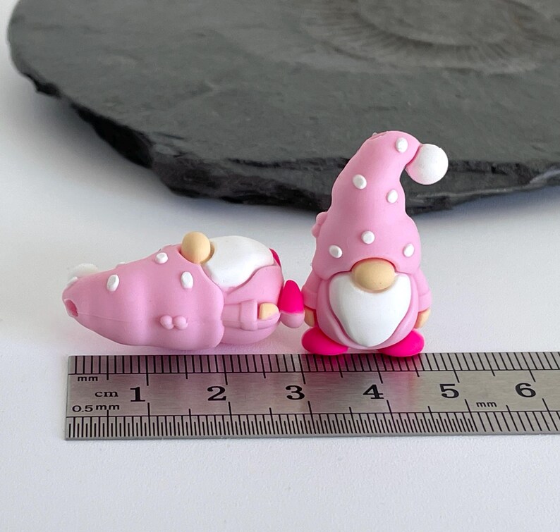 May include: Two pink gnome figurines with white polka dots and a ruler. One gnome is standing, the other is lying down. The standing gnome has a white beard and a pink hat with a white pom-pom. The ruler shows measurements in centimeters.
