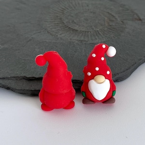 May include: Two red gnome figurines. One is plain, the other has white polka dots on its hat and a white beard. Both have a red hat with a white ball on top. The gnomes are on a white surface.