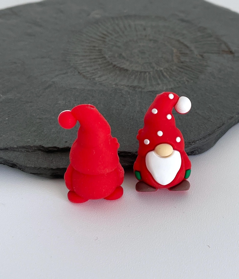 May include: Two red gnome figurines. One is plain, the other has white polka dots on its hat and a white beard. Both have a red hat with a white ball on top. The gnomes are on a white surface.