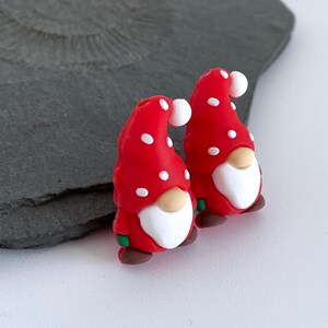May include: Two whimsical gnome figurines. Each gnome has a red hat with white polka dots, a white beard, a tan nose, and brown shoes. The gnomes have green accents and are set against a gray background.
