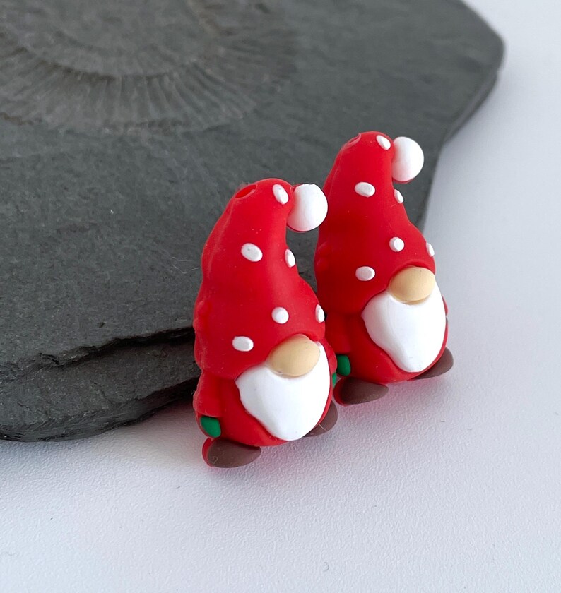 May include: Two whimsical gnome figurines. Each gnome has a red hat with white polka dots, a white beard, a tan nose, and brown shoes. The gnomes have green accents and are set against a gray background.