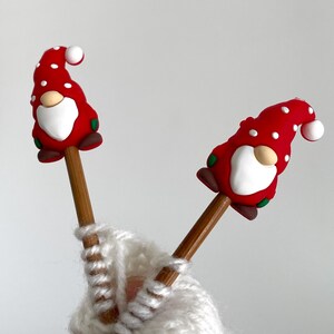 May include: Two wooden knitting needles topped with red gnome figures with white polka dots, white beards, and brown shoes. The needles are inserted into a white knitted item.