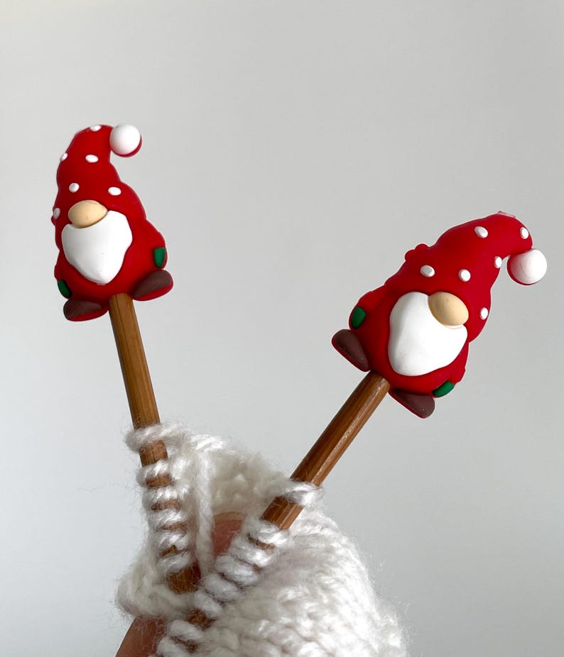 May include: Two wooden knitting needles topped with red gnome figures with white polka dots, white beards, and brown shoes. The needles are inserted into a white knitted item.