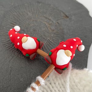 May include: Two red and white polka-dotted gnome-shaped knitting needle toppers. Each gnome has a white beard, tan nose, and a white ball on the tip of its hat. The toppers are attached to wooden knitting needles, with a white knitted item in the background.