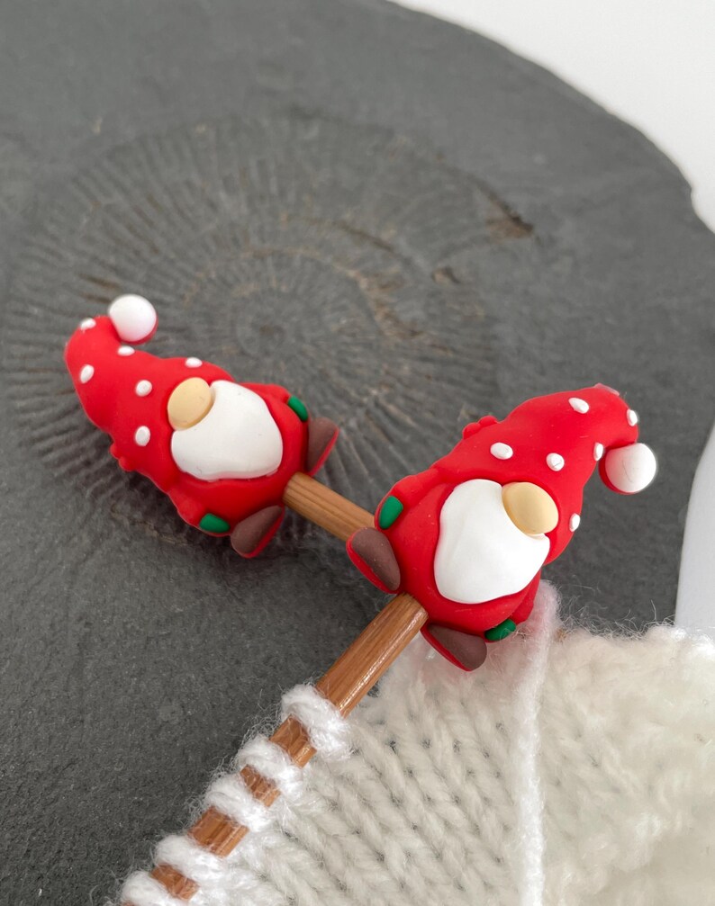 May include: Two red and white polka-dotted gnome-shaped knitting needle toppers. Each gnome has a white beard, tan nose, and a white ball on the tip of its hat. The toppers are attached to wooden knitting needles, with a white knitted item in the background.