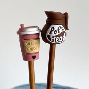 Può includere: Due puntali per ferri da maglia in legno. Uno è una tazza di caffè rosa con la scritta "BUT FIRST COFFEE" e un cuore nero. L'altro è una caffettiera marrone con la scritta "Pot Head".