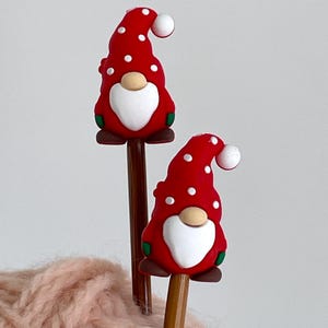 May include: Two pencils topped with red gnome figures. The gnomes have white polka dots, white beards, and a white ball on the end of their hats. The gnomes are on brown pencil shafts. The pencils are resting on a pink, fuzzy surface.