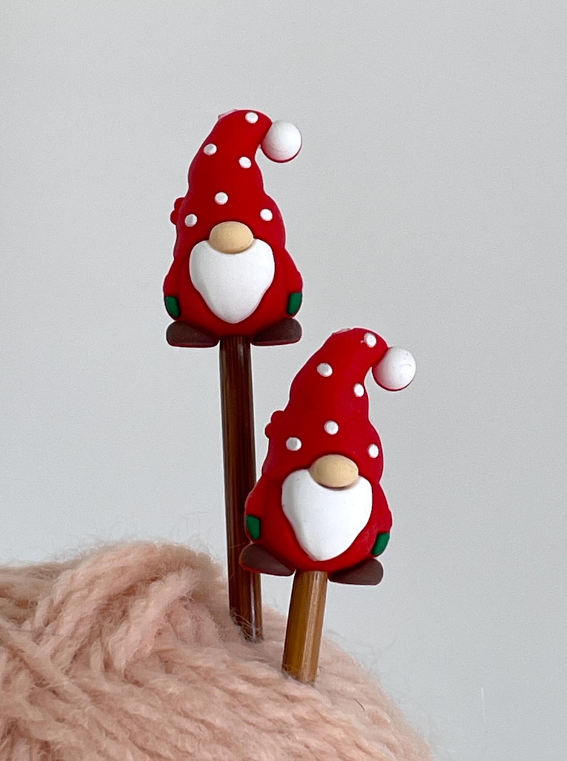 May include: Two pencils topped with red gnome figures. The gnomes have white polka dots, white beards, and a white ball on the end of their hats. The gnomes are on brown pencil shafts. The pencils are resting on a pink, fuzzy surface.