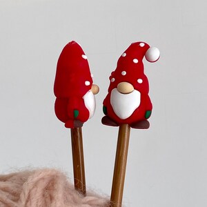 May include: Two Christmas gnome ornaments with red hats dotted with white spots. The gnomes have white beards, tan noses, and brown shoes. They are attached to wooden dowels, perfect for holiday decor.