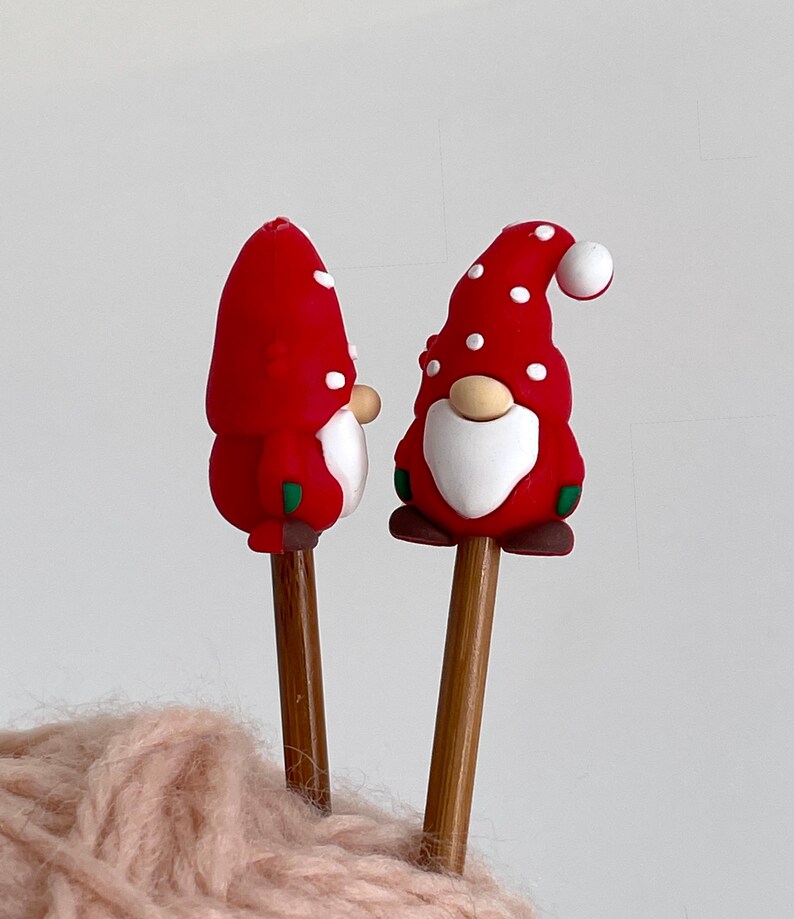 May include: Two Christmas gnome ornaments with red hats dotted with white spots. The gnomes have white beards, tan noses, and brown shoes. They are attached to wooden dowels, perfect for holiday decor.