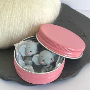 Pode incluir: Uma lata rosa com tampa a condizer, contendo duas estatuetas de elefantes cinzentos. Os elefantes têm orelhas grandes e bochechas rosadas. A lata está sobre uma superfície cinzenta escura, com uma bola de lã branca no fundo.
