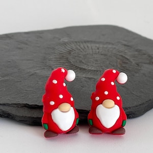 May include: Two Christmas gnome figurines. Each gnome has a red hat with white polka dots, a white beard, and a tan nose. The gnomes have green and brown accents. The figurines are on a dark gray surface.