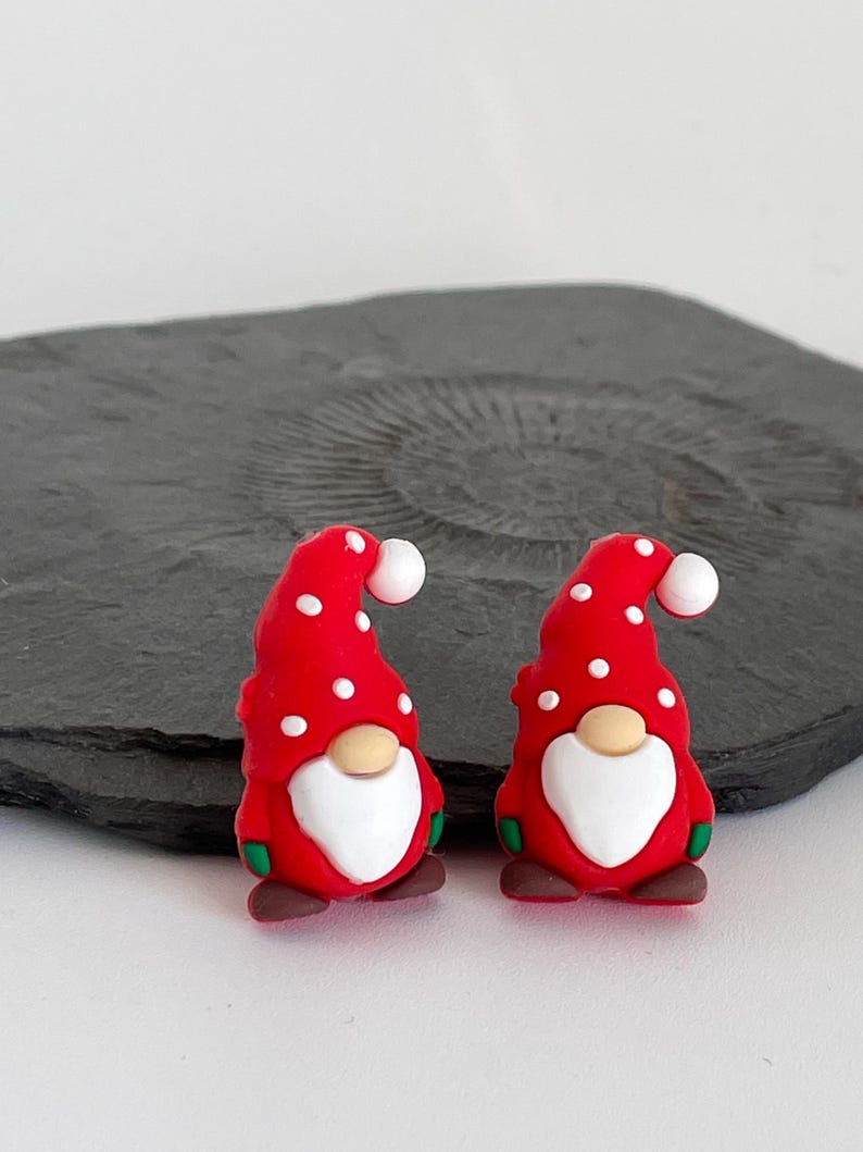 May include: Two Christmas gnome figurines. Each gnome has a red hat with white polka dots, a white beard, and a tan nose. The gnomes have green and brown accents. The figurines are on a dark gray surface.
