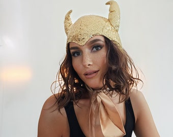 May include: A woman wearing a gold sequined devil horn headband with a nude satin scarf tied around her neck. She is wearing a black tank top.