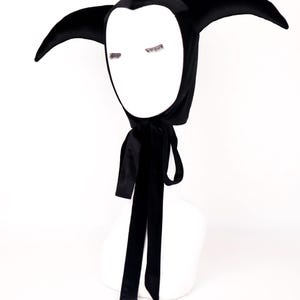 May include: A black velvet jester hood with long, pointed ears and a white face shape. The hood has a long, black satin ribbon tie. The hood is displayed on a white mannequin head.