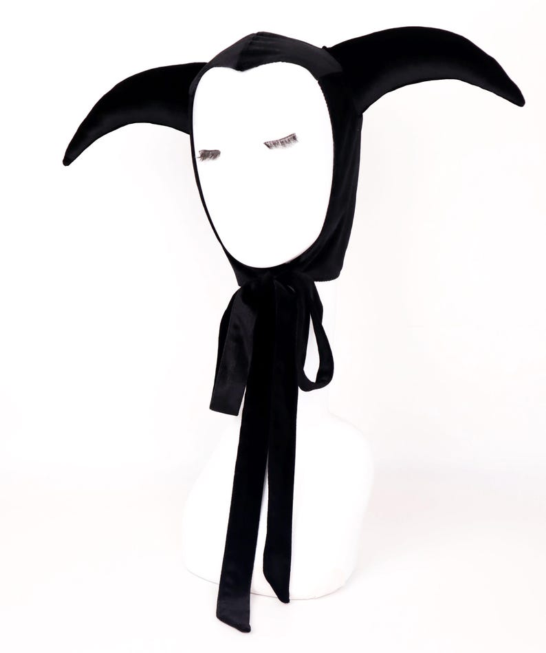 May include: A black velvet jester hood with long, pointed ears and a white face shape. The hood has a long, black satin ribbon tie. The hood is displayed on a white mannequin head.