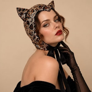 May include: A bonnet featuring a leopard print pattern covered in shimmering sequins with two pointed cat-like ears on top. The headpiece is secured under the chin with a smooth black satin ribbon tie that hangs down in two long, thin fabric straps.
