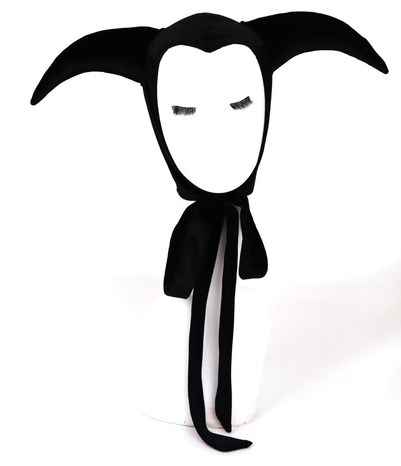 May include: A solid black velvet bonnet featuring two long, pointed, horn-like projections extending from the sides of the headpiece. The cap includes two long fabric ties that hang down from the chin area, securing the head covering in place.