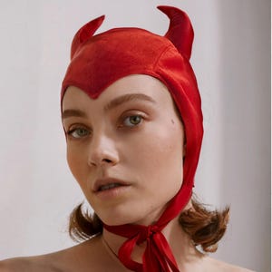 May include: A bright red velvet bonnet featuring two pointed horn-like structures on the top. The headpiece has a V-shaped front edge that sits on the forehead and includes long fabric ties that fasten securely under the chin with a bow.