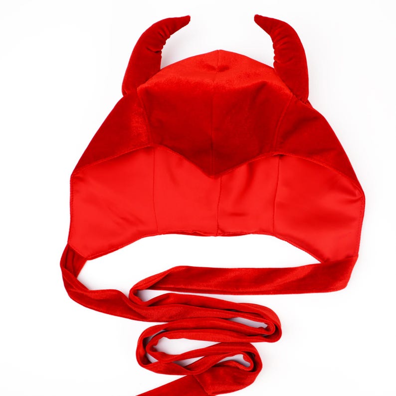 May include: A bright red velvet hood featuring two prominent, curved horns on top. The hood is constructed from a soft, plush fabric with a smooth, satin-like interior lining. Two long, matching red fabric ties extend from the base for fastening.