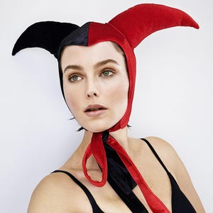 May include: A red and black velvet jester hat with a pointed tip and a long, flowing tie. The hat is worn by a person with short, dark hair.