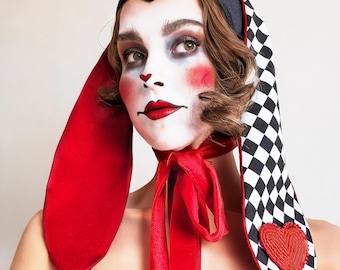 May include: A woman wearing a black and white checkered hat with red and white bunny ears. The hat has a red ribbon tied around the neck. The woman has red and white makeup on her face.