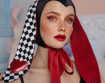 May include: A black and red bunny ear headband with a black and white checkered pattern and a red heart on one ear. The headband is worn by a person with red lipstick and a red ribbon tied around their neck.
