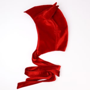May include: A bright red velvet hood featuring two pointed horn-like structures on the top. The hood is constructed from a soft, plush fabric and includes long, matching red velvet ribbon ties that extend from the base to secure the garment.