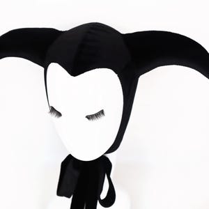 May include: A black velvet jester hat with three curved points. The hat is worn on a white mannequin head with closed eyes and attached eyelashes. A black scarf is draped around the neck.