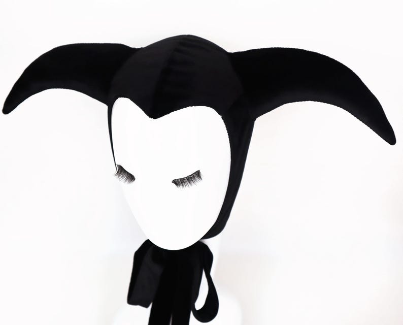 May include: A black velvet jester hat with three curved points. The hat is worn on a white mannequin head with closed eyes and attached eyelashes. A black scarf is draped around the neck.