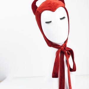 May include: A deep red velvet bonnet featuring two matching curved horns on the top. The headpiece has a pointed front edge that frames the forehead and includes long, integrated velvet ties that knot securely under the chin for a snug fit.