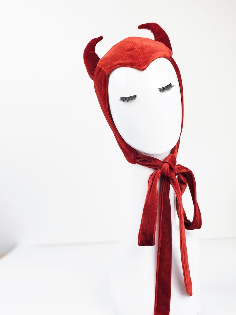 May include: A deep red velvet bonnet featuring two matching curved horns on the top. The headpiece has a pointed front edge that frames the forehead and includes long, integrated velvet ties that knot securely under the chin for a snug fit.