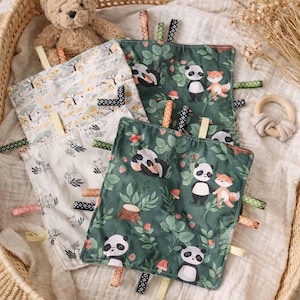 May include: Four baby tag blankets with various patterns, including pandas, foxes, and forest scenes. The blankets are square-shaped with colorful ribbons attached to the edges. One wooden teething ring is also visible.