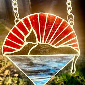 Könnte beinhalten: Eine Halskette mit Anhänger aus Buntglas mit einem Sonnenaufgangsdesign. Der Anhänger hat eine rot-weiße Sonne, eine schwarze Bergsilhouette und eine blau-weiße Wasserszene. Er hängt an einer silbernen Kette.