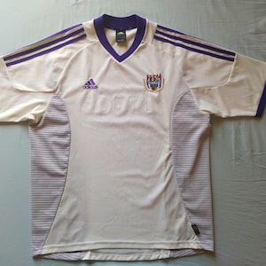 Puede incluir: Camiseta de fútbol crema con detalles morados. Presenta cuello en V, logotipo de Adidas y la palabra "FORTIS" en relieve. Las mangas tienen rayas moradas y la camiseta muestra un escudo del equipo.
