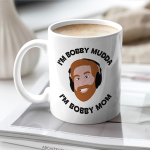May include: White ceramic coffee mug with a black and red design. The mug features a cartoon illustration of a man with a beard and headphones. The text on the mug reads "I'm Bobby Mudda I'm Bobby Mom".