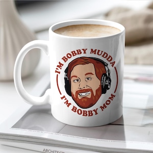 May include: White ceramic coffee mug with the text "I'M BOBBY MUDDA I'M BOBBY MOM" in a circular design around a cartoon illustration of a man with red hair and a beard wearing headphones. The mug is filled with a light brown liquid.
